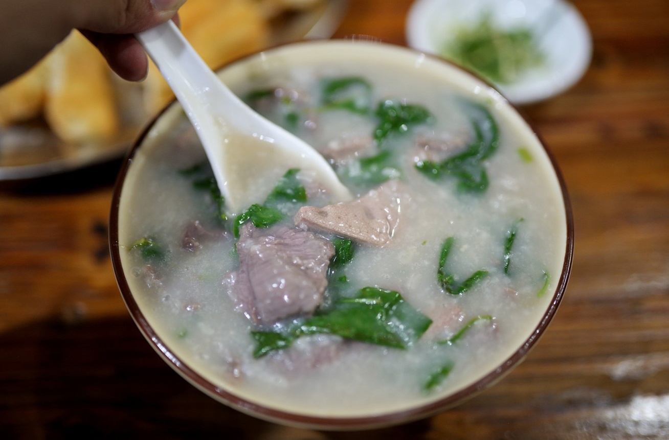  A large, steaming bowl of the Signature Pork Liver & Beef Congee, generously filled with slices of liver and beef, and garnished with green wolfberry leaves.