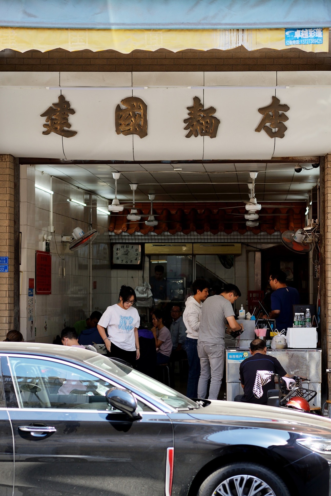  The humble, almost hidden storefront of Jianguo Noodle Shop, with a line of patient customers waiting outside on the narrow street, signaling its immense popularity.